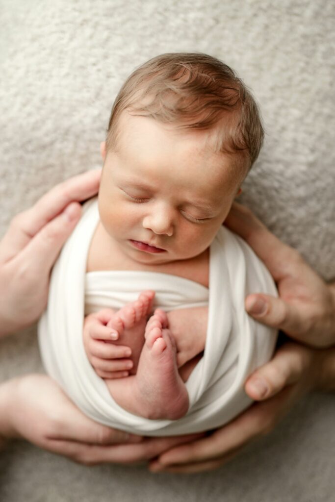 Newborn in neutral white wrap and background held in parents hands during Columbia, MD Photography Session