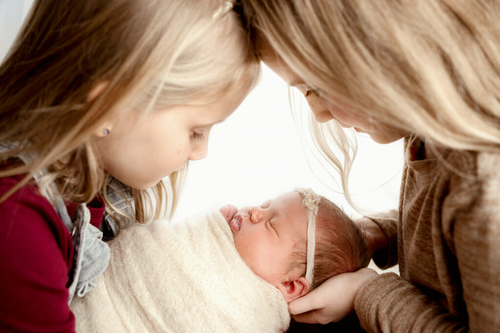 Newborn baby wrapped in white with two older sisters gazing over her in a triangle shape with a white backlit background