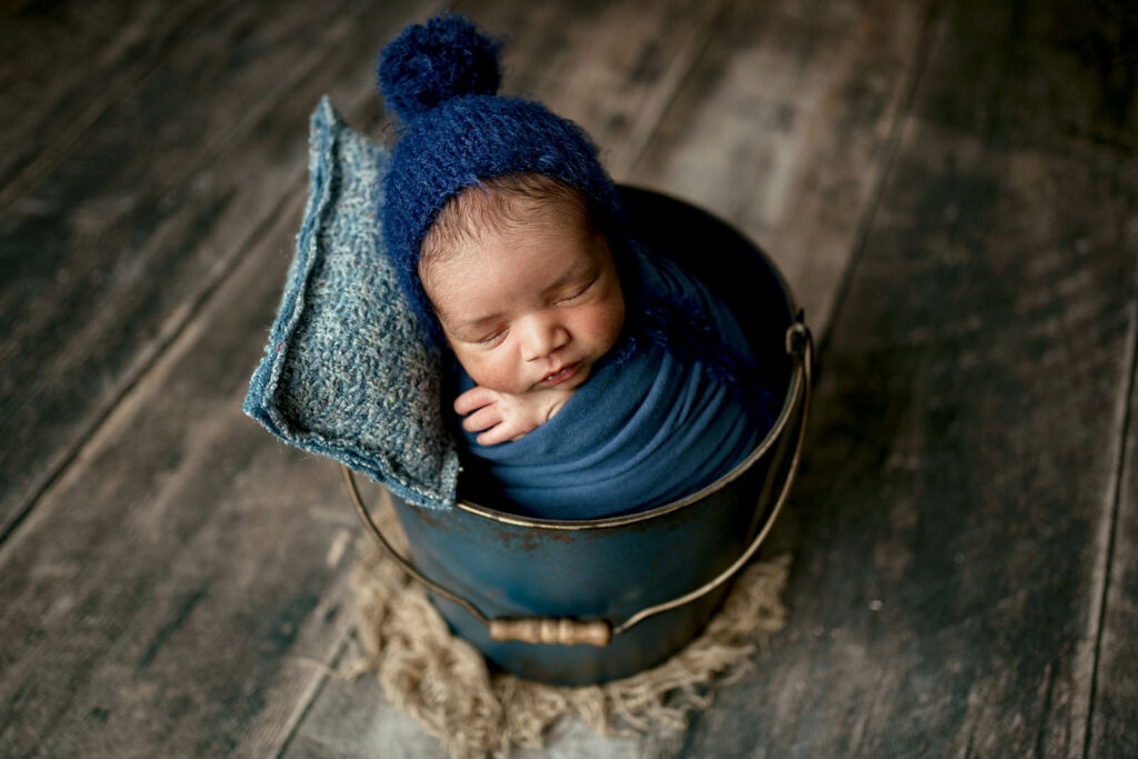 Baby boy wrapped in dark blue sweater with pom hat and matching pillow, in a blue bucket in his home in Reisterstown MD