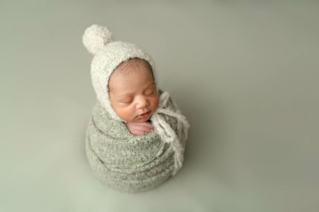 Sleeping baby wrapped in green sweater with winter bonnet on green backdrop