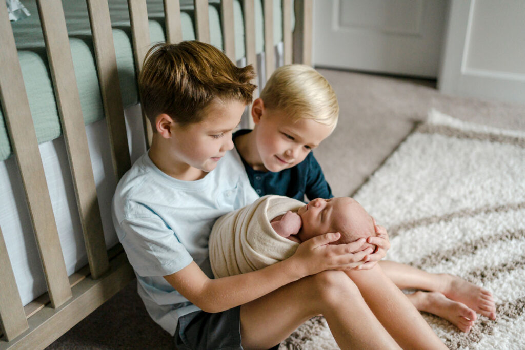 Two older brothers admire their newborn baby brother while holding him on the floor in front of his crib during a Lifestyle Newborn Photo Session in Howard County, MD