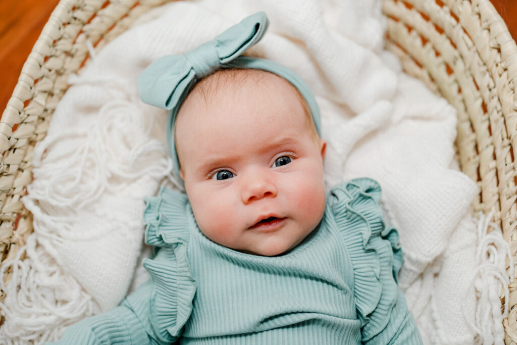 Baby in bright blue outfit matching her bright blue eyes looking at the camera while relaxing in a moses basket during her Lifestyle Newborn Photo Session in Baltimore MD