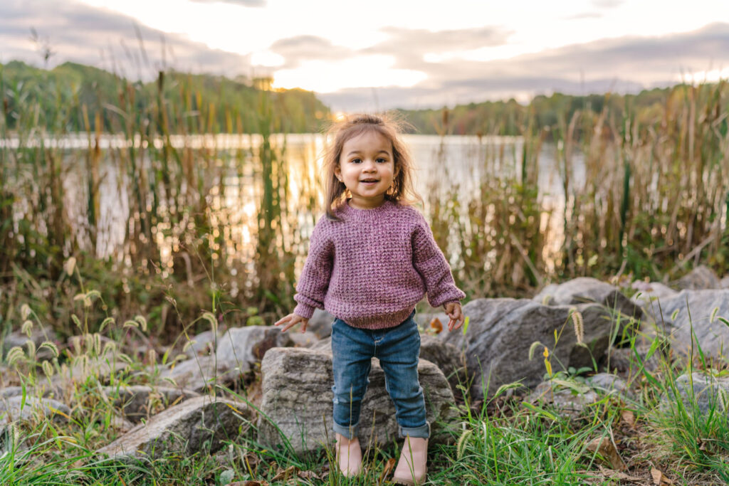 Toddler smiling while standing near rocks and pond grass in front of Centennial Lake at sunset.