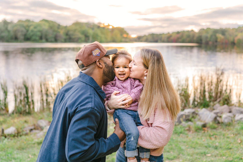 Parents kissing their toddlers cheeks as she smiles at the camera in front of Centennial lake at sunset.