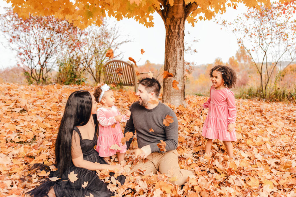 Family playing in bright orange leaves under a tree at the Howard County Conservancy