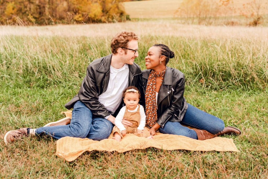 A family seated on a blanket in a picturesque field in Howard County MD. Parents are smiling at eachother while their baby sits between them looking at the camera