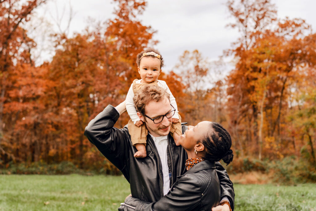 Baby sits on dad's shoulders while he and mom embrace as baby looks directly at the camera. Family photo session in front of lovely deep orange fall foliage in Howard County MD