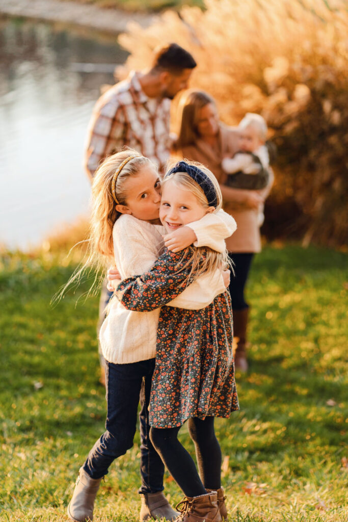 Sisters embrace while parents admire their baby sister in the background during their family's lifestyle photo session at sunset